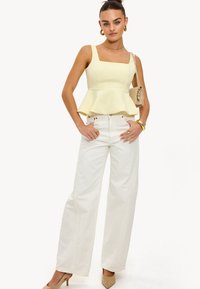 Haut peplum jaune avec décolleté carrée et bretelles larges, associé à un pantalon large blanc ; le mannequin est accessoirisé avec des bijoux en or et un sac.