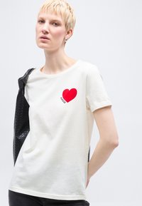 Γυναίκα με κοντά ξανθά μαλλιά φοράει λευκό μπλουζάκι με κόκκινη καρδιά και το κείμενο "ΑΜΟΥΡ", κρατώντας ένα μαύρο υφασμάτινο σακάκι πάνω σε έναν ώμο.