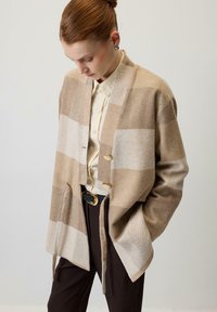 Bēšas krāsas vilnas kardigans ar platām rūtiņām, brīvs piegriezums, atvērts priekšpuses dizains, divas pogas un sānu sasienamie. Valkāts virs krēmkrāsas krekla un tumšām biksēm.