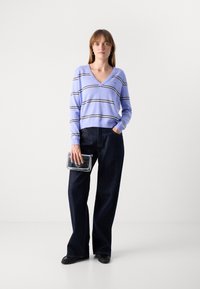 Pull violet clair à rayures horizontales, pantalon large en denim noir, tenant une pochette argentée, et chaussures plates noires.