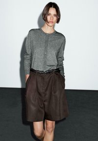 Cardigan gris à manches longues avec un devant à boutons, associé à un short plissé marron ; le modèle porte une ceinture noire tressée et a les mains dans les poches.