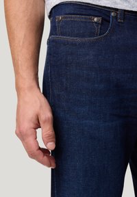 Man in een donkerblauwe denim jeans met zichtbare voorzak, met zijn hand ontspannen aan zijn zijde tegen een neutrale achtergrond.