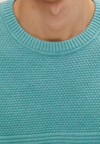 Personne portant un pull tricoté bleu clair texturé avec un col rond, montrant la partie inférieure du cou et le haut de la poitrine.
