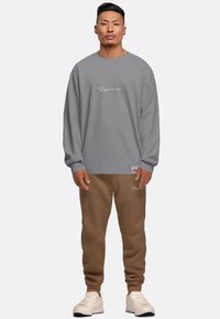 Felpa grigia a maniche lunghe con logo ricamato, abbinata a pantaloni della tuta marroni. Completa il look con sneakers bianche. Vestibilità rilassata, stile casual.