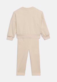 Μπεζ σετ δύο τεμαχίων με μακρυμάνικη μπλούζα που διαθέτει ριμπ μανσέτες και τελείωμα, συνδυασμένη με παντελόνι σε ίσια γραμμή, σε λευκό φόντο.