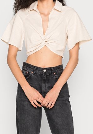 Kobieta w kremowym krótkim skręconym crop topie i czarnych dżinsach z wysokim stanem, ręce złożone z przodu na wysokości talii.