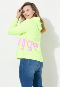 Neon groene hoodie met roze tekst op de achterkant, met een relaxed fit en een trekkoordkap, gecombineerd met blauwe spijkerbroeken.