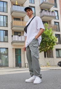 Hvit oversized t-skjorte, lys grønne cargo-bukser med lommer, og hvite sneakers. Svart caps og crossover-veske fullfører antrekket. Urban bakgrunn.