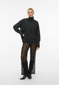 Přerostlý tmavě šedý rolák v kombinaci s černými krajkovými kalhotami s geometrickým vzorem a otevřenými boky. Špičaté černé kotníkové boty.