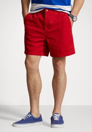 Shorts en coton rouge avec une coupe décontractée, dotés d'une fermeture à bouton et de poches latérales, associés à des baskets en toile bleue.