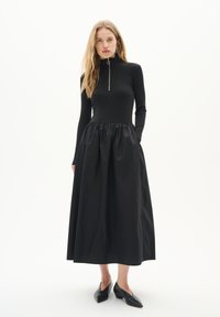 Rochie neagră cu un top strâmt din material cu dungi și fustă voluminoasă. Prezintă un guler înalt și detalii cu fermoar argintiu. Purtați cu pantofi negri ascuțiți.