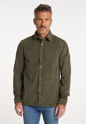 Mann mit kurzen grauen Haaren, der ein olivgrünes Cordhemd und blaue Jeans trägt und vor einem schlichten weißen Hintergrund steht.