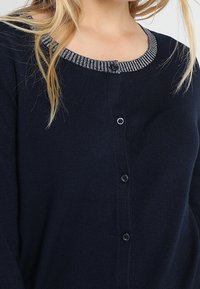 Žena v námořnickém svetrú s černými knoflíky a šedými pruhy kolem kulatého výstřihu, ukazující horní část trupu a vlasy po ramena v blond barvě.