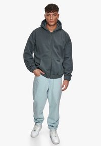 Mørkegrå zip-up hættetrøje med en afslappet pasform, forlommer og ribbede manchetter, parret med lyseblå bukser og hvide sneakers.