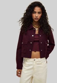 Burgundy cropped jacket with silver buttons and two front pockets, paired with a matching top and cream-colored jeans.