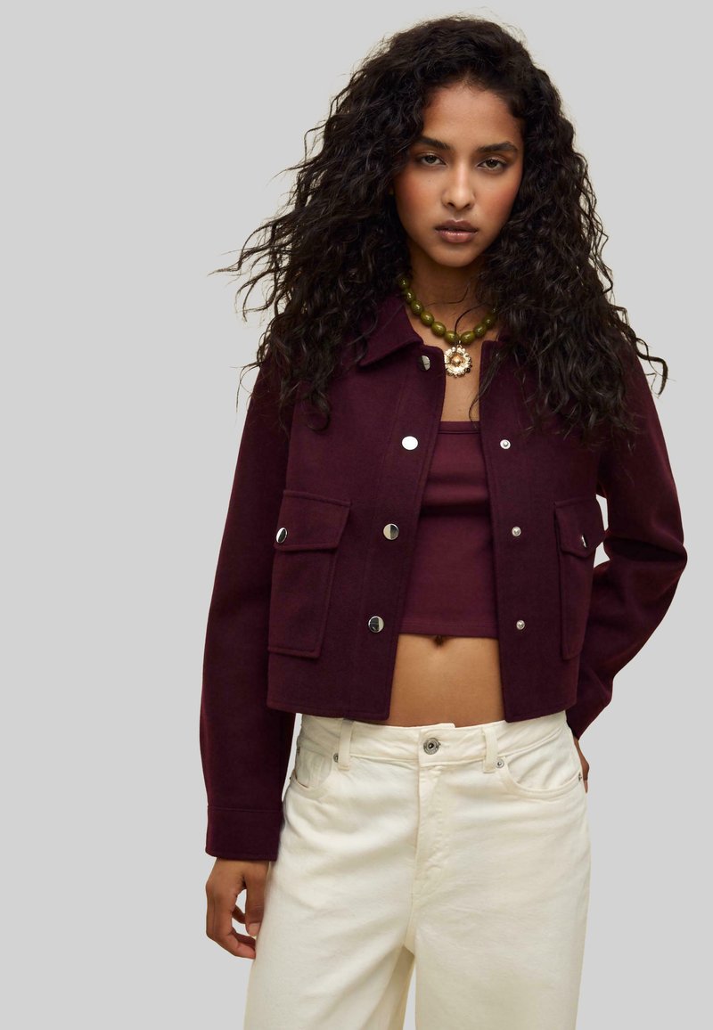 Burgundy cropped jacket with silver buttons and two front pockets, paired with a matching top and cream-colored jeans.