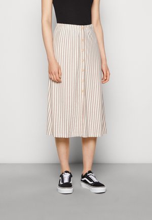 Vrouw die een midi-rok met verticale beige en witte strepen en knopen en een zwarte mouwloze top draagt, gecombineerd met zwart-witte sneakers.