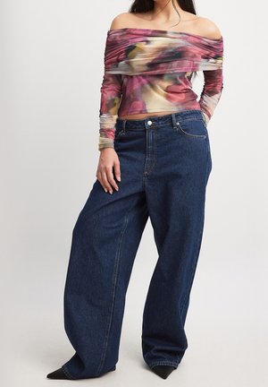 Femme portant un jean bleu foncé à jambes larges et un haut à manches longues, épaules dénudées, avec un motif abstrait coloré.