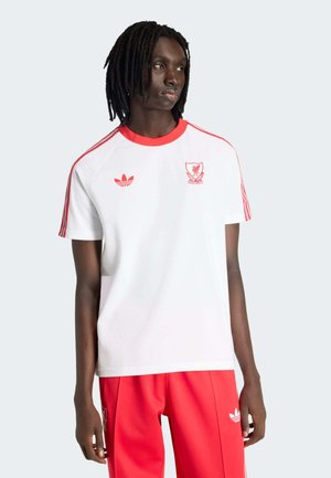 Young man with medium-length dreadlocks wearing white and red Liverpool football club Adidas shirt and red track pants, looking left.