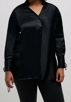 Personne portant un chemisier ample en satin noir à manches longues et un pantalon noir, debout devant un fond clair uni.