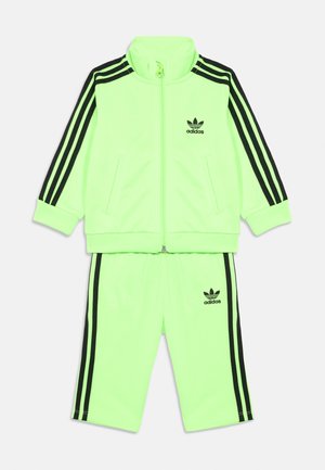 Neon zelena Adidas trenirka s črnimi tridelnimi trakovi na rokavih in hlačah, ki vključuje zadrgo na jakni in logotip na prsih ter stegnu.