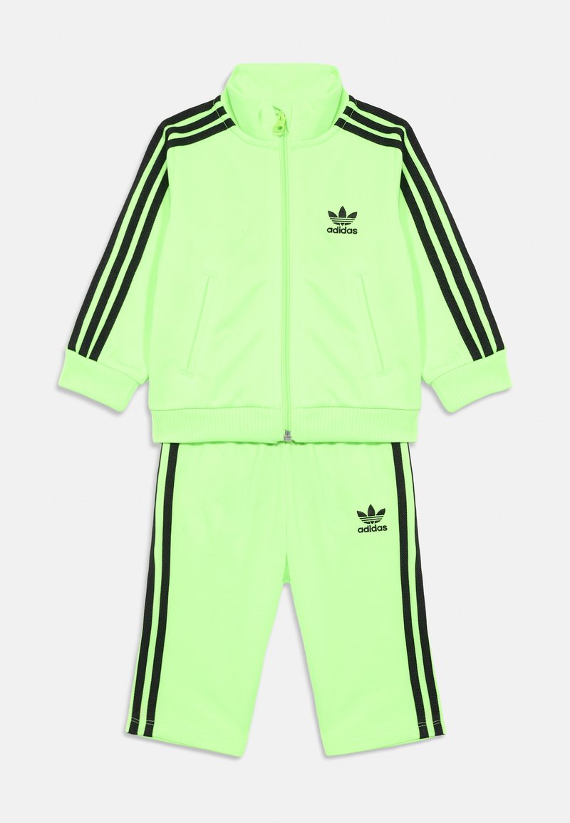 Neon zöld Adidas melegítő szett fekete háromsávos részletekkel az ujjakon és a nadrágon, cipzáras dzsekivel és emblémával a mellkason és a combon.