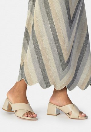 Pies con sandalias beige de tacón bloque con tiras cruzadas, bajo una falda con borde ondulado y rayas diagonales en tonos neutros.