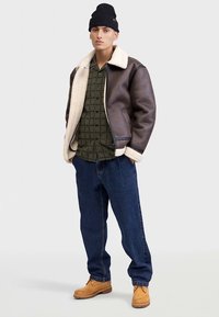 Jachetă bomber maro din piele cu căptușeală din blană, cămașă verde cu model, blugi albaștri și cizme maro deschis. Căciula neagră completează ținuta.