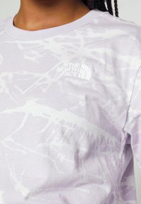 Jasnofioletowa koszulka z krótkim rękawem z białym abstrakcyjnym wzorem i białym logo The North Face na piersi. Tkanina bawełniana.