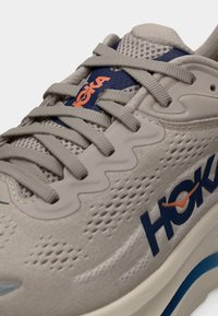 Κοντινό πλάνο σε μπεζ αθλητικό παπούτσι Hoka με γκρι κορδόνια, λογότυπο σε μπλε και πορτοκαλί, επάνω μέρος από δίχτυ και μαλακή σόλα με μπλε λεπτομέρεια.