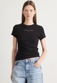 Melna kokvilnas T-kreklu ar īsām piedurknēm, uz krūtīm redzams spilgti baltā krāsā "TOMMY JEANS" logo. Apvienots ar gaiši ziliem, augsto jostasvietu džinsiem.