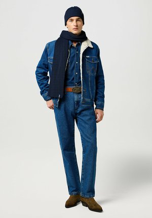 Jeansjack met schapenvachtkraag, blauwe overhemd, blauwe spijkerbroek, marineblauwe gebreide beanie, en sjaal, bruine riem, en bruine hakken.