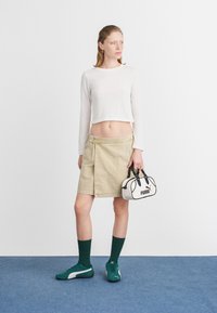 Fiatal nő áll a kék padlón, fehér, hosszú ujjú cropped topot, bézs kötött szoknyát, zöld zoknit és sportcipőt visel, miközben egy kis fehér Puma táskát tart a kezében.
