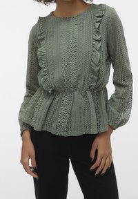 Top in pizzo di verde tenue con bordi arricciati, motivi testurizzati, maniche lunghe e vita peplum. Presenta elaborati disegni a intaglio.
