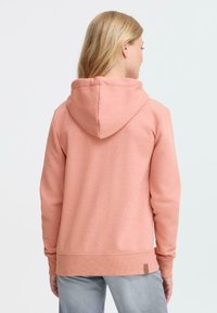 Sweat à capuche rose avec une poche kangourou, un cordon de serrage ajustable, des poignets et un ourlet côtelés, et une étiquette marron sur le côté. Texture douce.