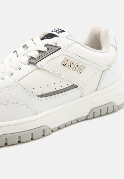 White sneakers with a leather and textile upper, featuring grey accents, perforated sides, and a logo detailing on the side panel.