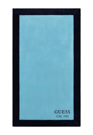 Tapis en peluche turquoise avec une bordure bleu marine foncé, comportant une étiquette noire imprimée indiquant "GUESS CAL. 1981" dans le coin inférieur droit.