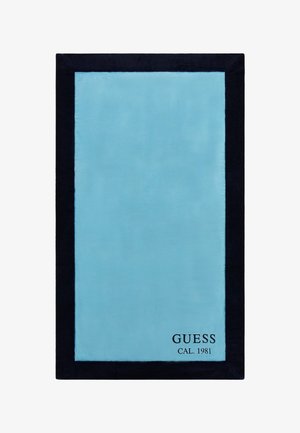 Turkusowy pluszowy dywan z ciemnogranatowym wykończeniem, z czarną etykietą z nadrukiem "GUESS CAL. 1981" w prawym dolnym rogu.