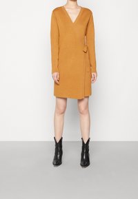 Abito avvolgente senape con maniche lunghe, scollatura a V e dettaglio con cinturino laterale. Indossato con stivaletti neri alla caviglia, che mostrano un design elegante e una texture morbida.