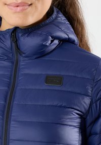Marinblå pufferjacka med huva, med en frontdragkedja, horisontell kviltad struktur och en liten svart logotags på bröstet.