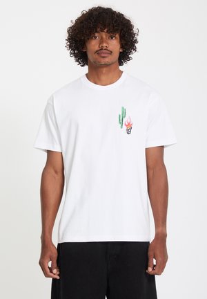Homme aux cheveux bouclés portant un t-shirt blanc avec un petit dessin vert de cactus et une flamme rose sur le côté gauche de la poitrine, associé à un pantalon noir.
