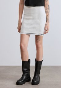 Λευκή μίνι φούστα ριμπ πλεκτή με οριζόντια ρίγα στο κάτω μέρος, συνδυασμένη με μαύρες μπότες μέχρι το γόνατο. Ορατό τατουάζ στο μηρό.