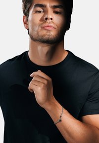 Silver chain bracelet on wrist, featuring a small charm. Black fitted t-shirt with a round neckline, plain design, smooth texture.