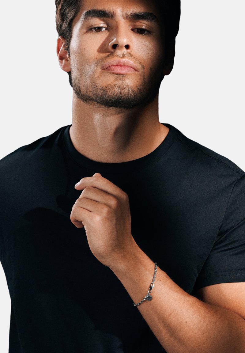 Silver chain bracelet on wrist, featuring a small charm. Black fitted t-shirt with a round neckline, plain design, smooth texture.