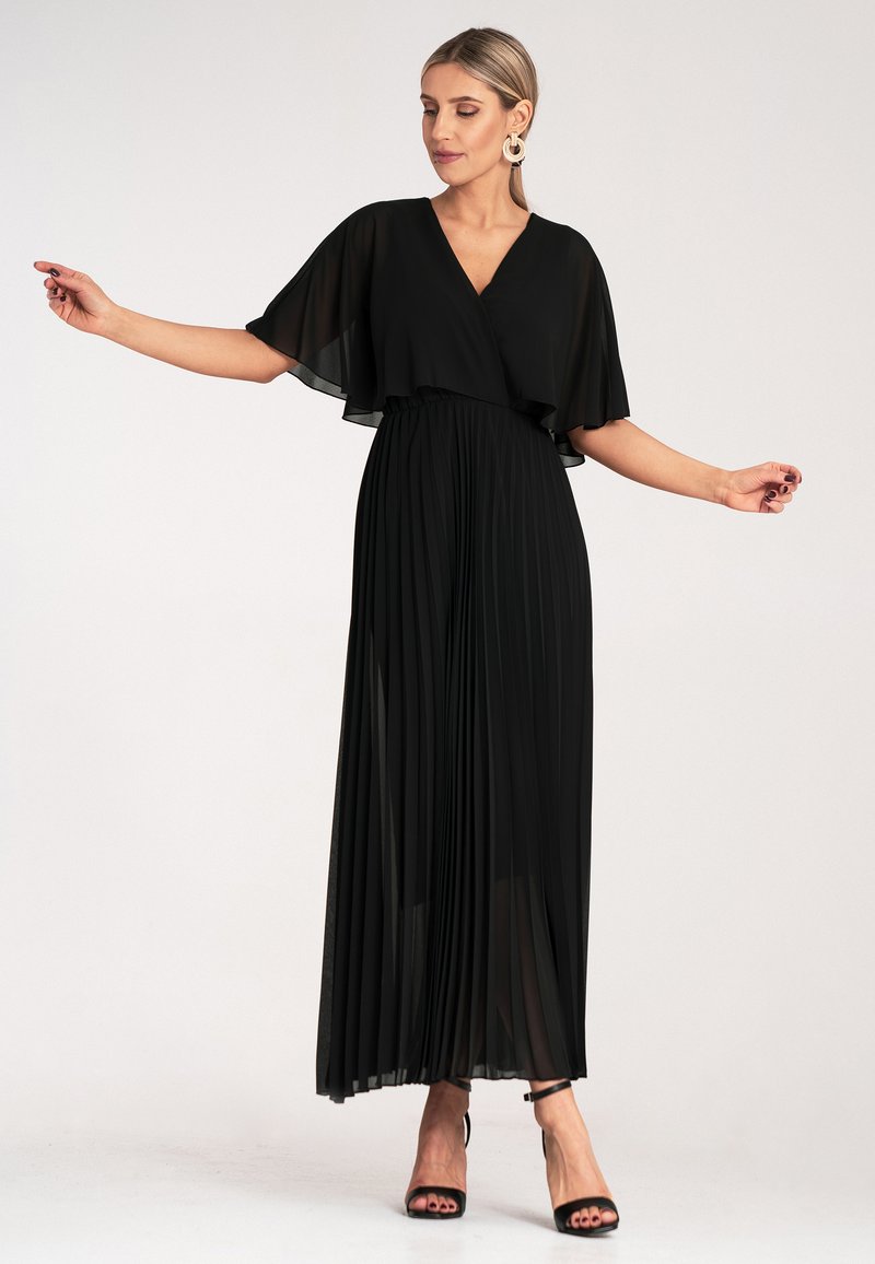 Czarna, plisowana sukienka maxi z dekoltem w serek i rękawami-czapami. Wykonana z lekkiego materiału. Modelka ma na sobie czarne sandały na wysokim obcasie i wiszące kolczyki.