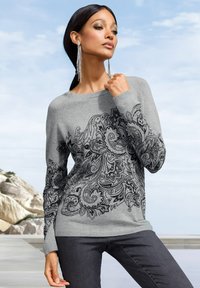 Frau in einem grauen Pullover mit schwarzem floralen Paisleymuster, dunkler Hose und langen silbernen Ohrringen vor einem bewölkten Himmel und einer felsigen Kulisse.