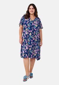Robe à imprimé floral avec un fond marine et des fleurs roses et bleues, manches courtes, décolleté en V et une silhouette décontractée. Associée à des chaussures bleues.