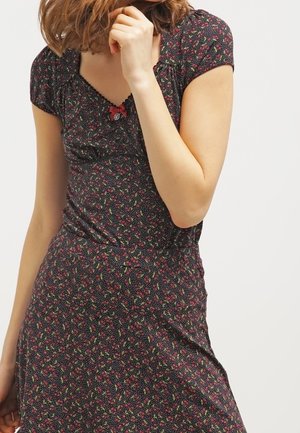 Kobieta w krótkiej sukience na krótki rękaw, czarnej z małymi czerwonymi kwiatowymi wzorami i dekoltem w serek z guziczkami, pozująca z ręką przy twarzy.