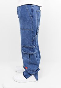Hellblaues Denim-Cargo-Hose mit mehreren Taschen, lockerer Passform und einem roten Logo-Patch. Gestylt mit weißen Sneakers.