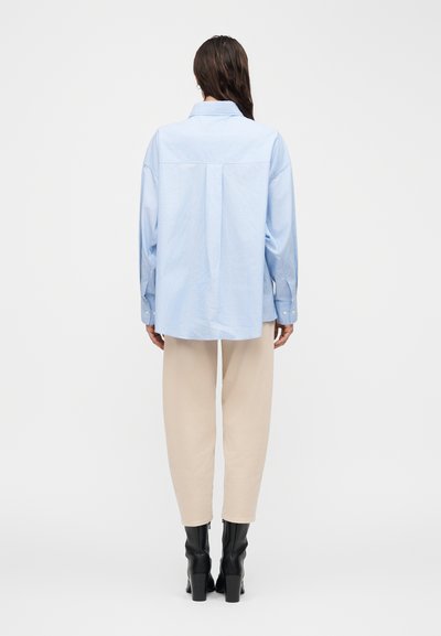 Γυναίκα που στέκεται με την πλάτη γυρισμένη, φοράει ανοιχτό μπλε φαρδιά πουκάμισο, μπεζ παντελόνι και μαύρες μπότες με τακούνι σε άσπρο φόντο.